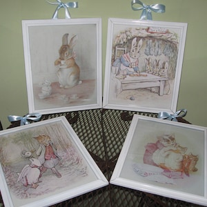 May include: Four white framed pictures with blue ribbon accents. Each picture features a different illustration of Peter Rabbit and his friends. The first picture shows Peter Rabbit holding a teacup. The second picture shows Peter Rabbit sitting at a table with other rabbits. The third picture shows Peter Rabbit and a duck. The fourth picture shows Peter Rabbit and a cat.