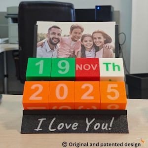 Op de afbeelding: Een kleurrijke, herbruikbare kalender met de datum 19 november 2025. De datum wordt weergegeven op groene, rode, oranje en witte blokken, met de tekst "I Love You!" op een zwarte basis. Inclusief een familiefoto.