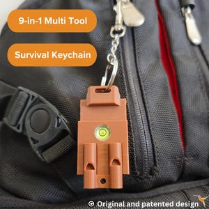 Puede incluir: Un llavero de supervivencia marrón multiherramienta 9 en 1. El llavero incluye un nivel, dos silbatos y un llavero. El texto "9-in-1 Multi Tool" y "Survival Keychain" se muestra en la imagen. El llavero está sujeto a una mochila negra.