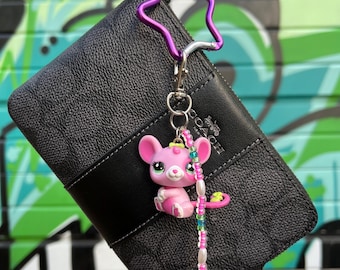 Op de afbeelding: Een zwarte Coach-portemonnee met een roze en witte kralen sleutelhanger met een klein roze cartoondier met groene ogen. De sleutelhanger is bevestigd aan een paarse stervormige metalen haak.