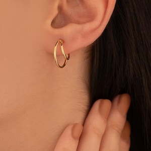 Puede incluir: Pendientes de aro dorados. Cada pendiente presenta un diseño de doble capa, con dos aros circulares conectados. La imagen muestra una vista de cerca de la oreja y las joyas.