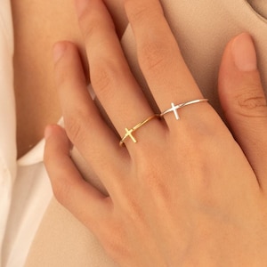 Könnte beinhalten: Zwei zarte Ringe aus Gold und Silber mit einem Kreuzdesign. Die Ringe werden am Ringfinger einer Hand getragen.