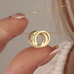 May include: A gold-colored necklace featuring three interlocking rings. Each ring has engraved text. The necklace is held by a hand, with the chain extending towards the neck. The background is a soft, neutral color.