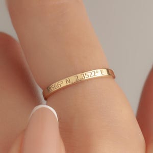 Puede incluir: Un anillo dorado con coordenadas grabadas se lleva en un dedo. El anillo es delgado y tiene un diseño sencillo. El fondo es de un color suave y neutro. El anillo es una joya.