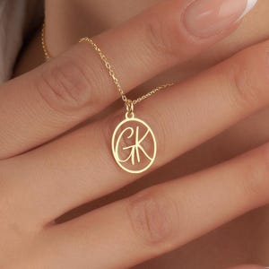 May include: A gold-colored necklace with a delicate chain and a circular pendant. The pendant features the intertwined letters "GK" in a stylized script. The necklace is worn around the neck.