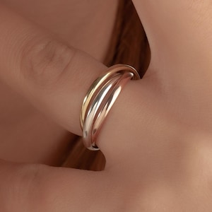 May include: A tri-colour ring with three interwoven bands, in gold, silver, and rose gold. The ring is worn on a finger, highlighting its elegant design and smooth finish. A close-up shot shows the ring's details.