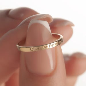 以下が含まれることがあります： 「Olivia」と「Penelope」の名前が刻まれたゴールドカラーのリング。ハートのシンボルで区切られています。白い背景にリングが掲げられています。