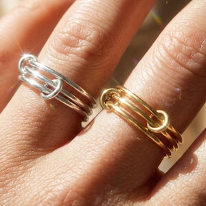 May include: Two rings, one silver and one gold, are displayed on a hand. Each ring features a unique design with three parallel bands connected by a small, curved element. The rings are simple yet elegant, suitable for everyday wear.