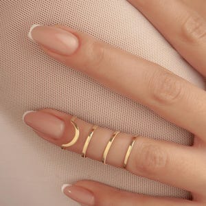 May include: A hand adorned with multiple gold rings. The rings are delicate and minimalist, including a crescent moon design. The nails are painted with a pale pink hue. The rings are simple bands. The image is a close-up.