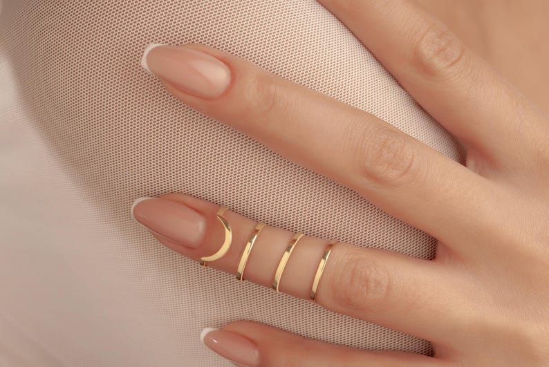 May include: A hand adorned with multiple gold rings. The rings are delicate and minimalist, including a crescent moon design. The nails are painted with a pale pink hue. The rings are simple bands. The image is a close-up.