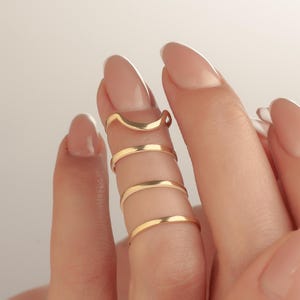 May include: Close-up of gold-coloured finger rings. The rings are worn on multiple fingers, with a curved ring at the top and several bands below. The rings are made of a shiny, metallic material.