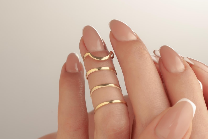 May include: Close-up of gold-coloured finger rings. The rings are worn on multiple fingers, with a curved ring at the top and several bands below. The rings are made of a shiny, metallic material.