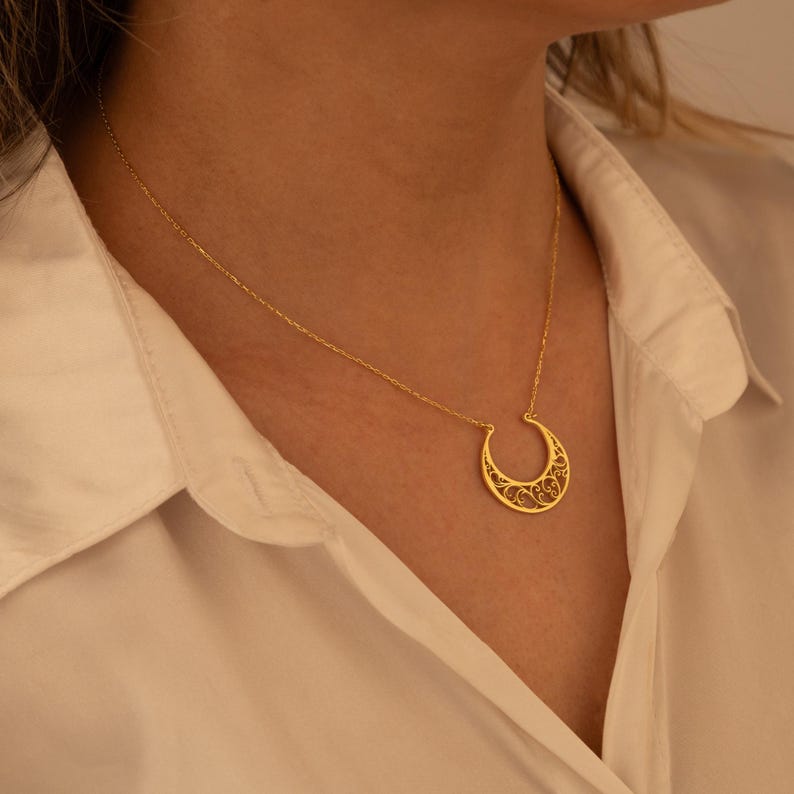May include: A gold-colored necklace featuring a crescent moon pendant with an intricate, openwork design. The necklace is worn against a white collared shirt, highlighting the jewelry's delicate details and warm hue.