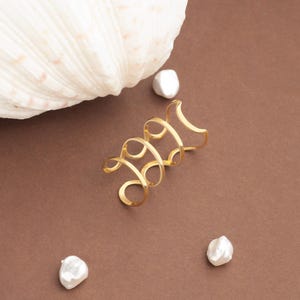 May include: A gold ear cuff with a spiral design, on a brown surface. The ear cuff is next to a large white seashell and three white, irregular-shaped pearls.