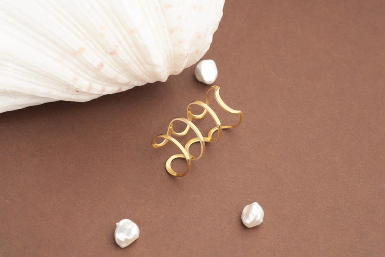 May include: A gold ear cuff with a spiral design, on a brown surface. The ear cuff is next to a large white seashell and three white, irregular-shaped pearls.