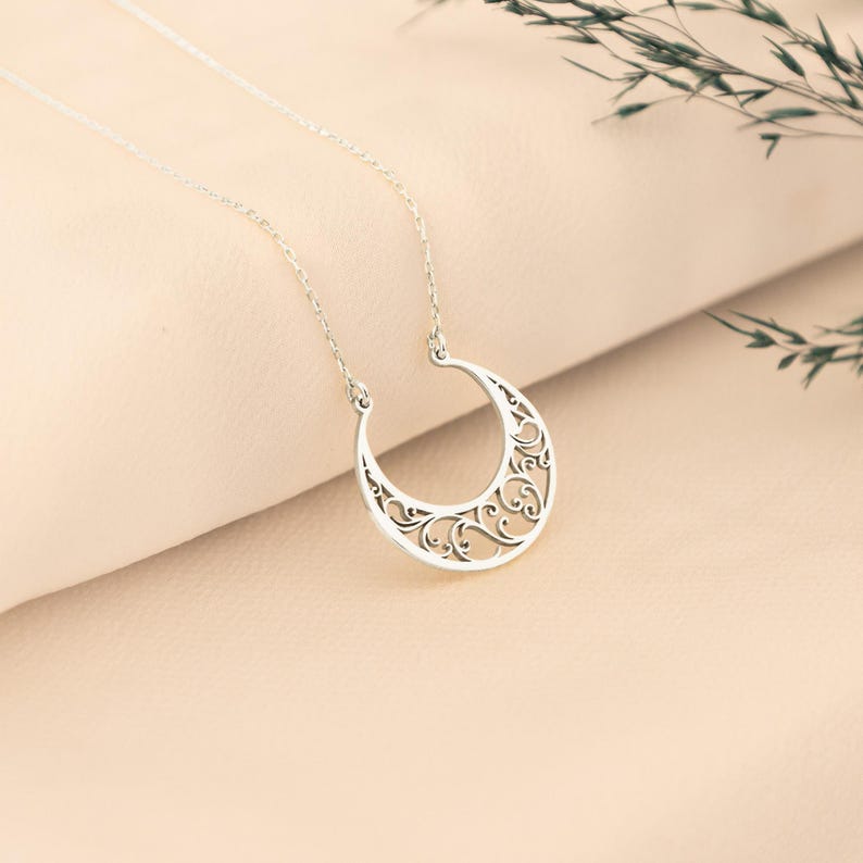 May include: A silver-colored crescent moon pendant necklace. The pendant features an openwork design with intricate swirling patterns. The necklace chain is delicate and attached to the top of the moon shape. The background is a soft, neutral color.