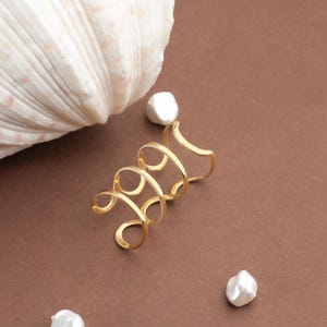 May include: A gold ear cuff in a spiral design sits on a brown surface, next to a large white seashell and three white pearls. The ear cuff has an open design. The pearls are irregularly shaped.