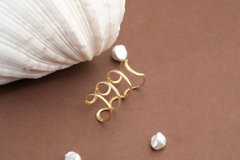 May include: A gold ear cuff in a spiral design sits on a brown surface, next to a large white seashell and three white pearls. The ear cuff has an open design. The pearls are irregularly shaped.