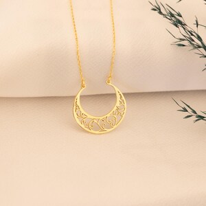 May include: A gold-colored crescent moon pendant necklace. The moon pendant features an open filigree design with swirling patterns. The necklace hangs on a delicate gold chain, displayed against a soft, off-white background.