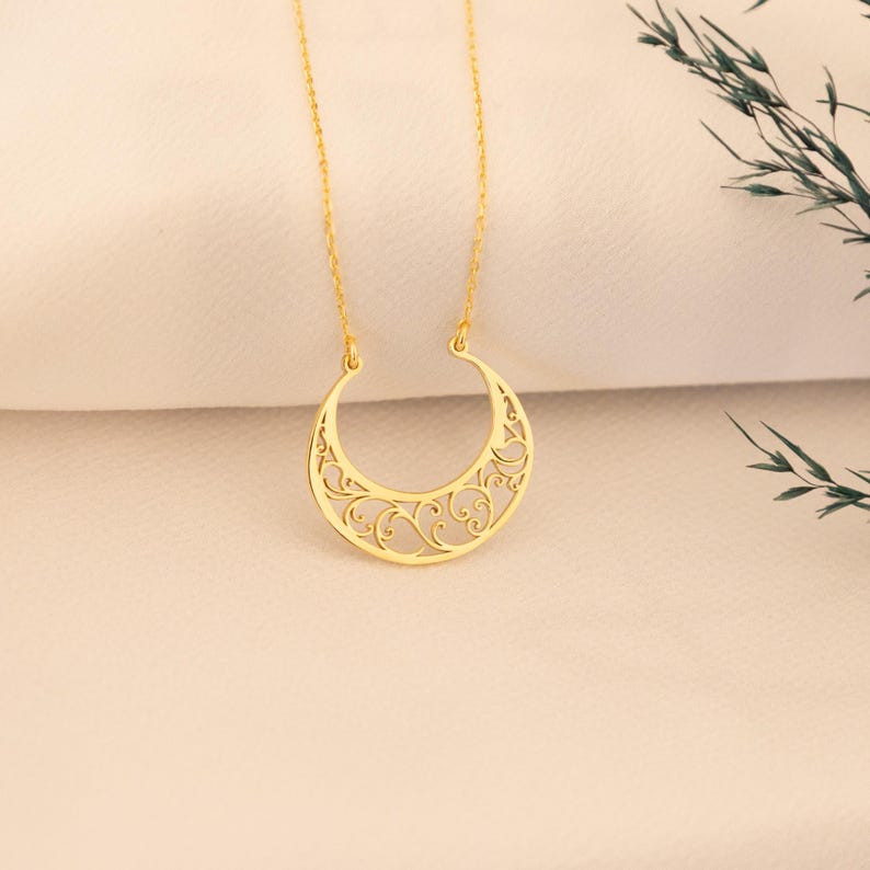 May include: A gold-colored crescent moon pendant necklace. The moon pendant features an open filigree design with swirling patterns. The necklace hangs on a delicate gold chain, displayed against a soft, off-white background.