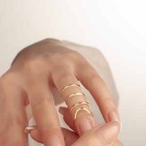 May include: A hand displays four delicate, gold-coloured rings. The rings are thin and have a wavy design, creating a unique and elegant look. The rings are worn on the index and middle fingers.