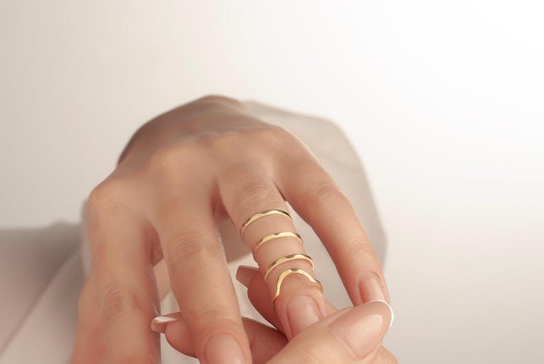 May include: A hand displays four delicate, gold-coloured rings. The rings are thin and have a wavy design, creating a unique and elegant look. The rings are worn on the index and middle fingers.