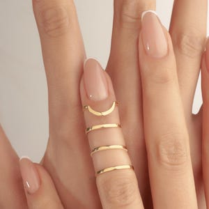 May include: Close-up of a hand adorned with gold rings. The rings include a crescent moon shape and three thin bands. The nails are painted with a light pink colour and have white tips.