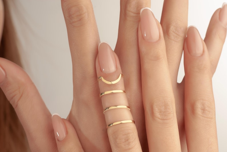 May include: Close-up of a hand adorned with gold rings. The rings include a crescent moon shape and three thin bands. The nails are painted with a light pink colour and have white tips.