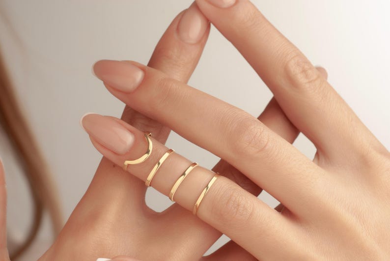 May include: A hand adorned with several gold rings. The rings are delicate and minimalist, with a crescent moon design on the top finger. The other rings are plain bands. The rings are on a light skin tone.