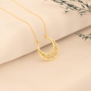 May include: A gold-colored necklace featuring a crescent moon pendant with an intricate, openwork design. The pendant hangs from a delicate chain. The necklace is displayed on a cream-colored surface, with a sprig of greenery in the background.