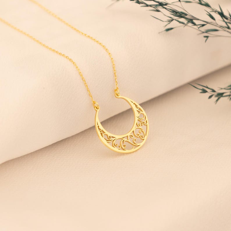 May include: A gold-colored necklace featuring a crescent moon pendant with an intricate, openwork design. The pendant hangs from a delicate chain. The necklace is displayed on a cream-colored surface, with a sprig of greenery in the background.