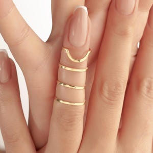 May include: Gold-coloured finger rings in various styles. The rings include a curved top ring and several thin, spiral rings. The rings are worn on a finger with a light-coloured nail polish.
