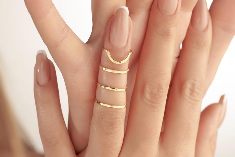 May include: Gold-coloured finger rings in various styles. The rings include a curved top ring and several thin, spiral rings. The rings are worn on a finger with a light-coloured nail polish.