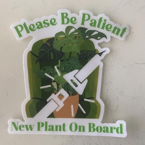 May include: A white sticker with green text that reads "Please Be Patient New Plant On Board". The sticker features a cartoon illustration of a potted plant with a seatbelt on.