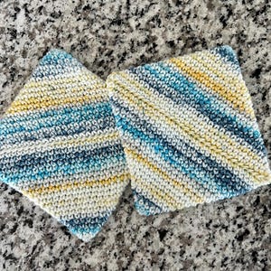 Op de afbeelding: Twee vierkante gehaakte onderzetters met een diagonaal streeppatroon in tinten blauwgroen, marineblauw, geel en crème. Het garen heeft een getextureerd, gemêleerd uiterlijk met een consistent steekpatroon over beide vierkante stoffen delen.