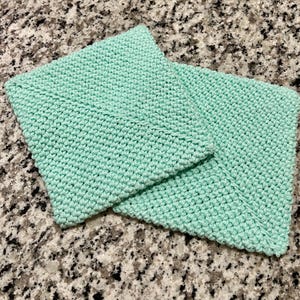 May include: Two square, mint green crocheted washcloths featuring a dense, textured stitch pattern. The items are folded and layered, displaying a consistent, uniform weave throughout the fabric with clean, finished edges on all sides.