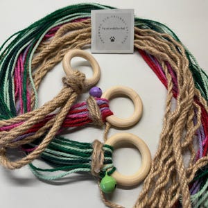 May include: A cat toy made with natural materials. It features three wooden rings with colourful yarn and jute rope. The toy is handmade and eco-friendly. The tag reads "Handmade Eco-Friendly Cat Toys".