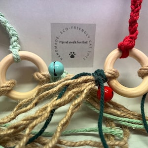 May include: Handmade eco-friendly cat toy with wooden rings, jute, and colourful cords. The toy features bells and tassels. The label reads "Handmade Eco-Friendly Cat Toys My cat would love that!"