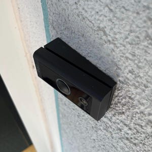 Könnte beinhalten: Eine schwarze, rechteckige Türklingelkamera, montiert an einer strukturierten, hellgrauen Wand. Die Türklingel hat eine kreisförmige Linse und eine kleinere Linse darunter. Der Markenname "Ring" ist sichtbar.