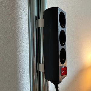 Könnte beinhalten: Eine schwarze Steckdosenleiste mit drei Steckdosen und einem roten Ein-/Ausschalter ist mit weißen Clips an einer silbernen Stange befestigt. Die Steckdosenleiste hat ein schwarzes Kabel und ist eingesteckt.