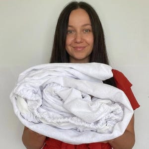 以下が含まれることがあります： 控えめな花柄の白いフィットシーツが丸められています。赤いドレスを着た人がシーツを持っています。背景はプレーンな白い壁です。