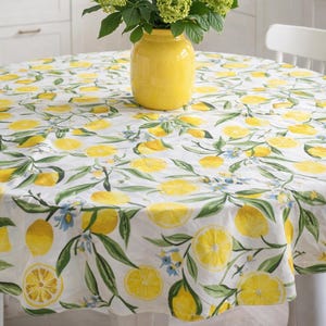 May include: A round tablecloth with a lemon pattern, featuring yellow lemons and green leaves on a white background. A yellow vase holds green flowers, and a yellow kettle and bowl are visible in the background. The tablecloth is approximately 60 inches in diameter.