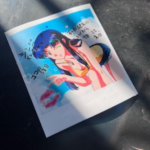May include: A colorful anime-style illustration on a white background. The artwork depicts a woman in a yellow bikini and denim shorts, with long dark hair. Japanese text and a kiss mark are also visible.