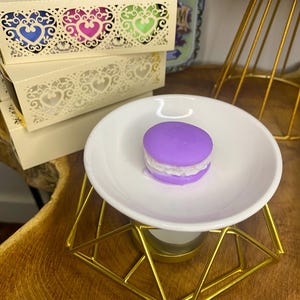 Pode incluir: Um derretedor de cera em forma de macaron roxo está em um prato branco sobre um suporte geométrico dourado. Atrás, caixas creme com recortes em forma de coração em azul, rosa e verde. As caixas estão rotuladas como "BOMBONES".