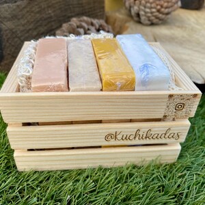 Puede incluir: Una caja de madera con cuatro jabones en tonos rosa, beige, amarillo y azul. La caja tiene grabado el nombre de Instagram "@Kuchikadas". La caja está sobre hierba verde.