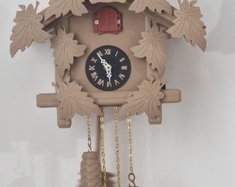 Könnte beinhalten: Eine beige Kuckucksuhr mit einer Hirschkopf-Schnitzerei oben. Die Uhr hat blattförmige Akzente, ein schwarzes Zifferblatt mit weißen römischen Ziffern und zwei tannenzapfenförmige Gewichte, die an Ketten hängen.