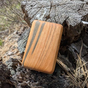 May include: A rectangular wooden object with rounded corners, featuring a light brown base with dark gray streaks. The wood grain is visible. The object is resting on a weathered, textured wooden surface, with a natural outdoor background.