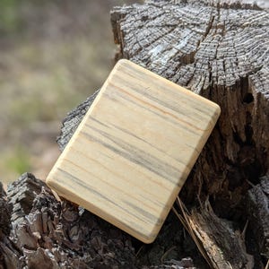 Puede incluir: Un objeto rectangular de madera con esquinas redondeadas, de color beige claro con vetas grises oscuras. La veta de la madera es visible. Descansa sobre un tocón de árbol desgastado y texturizado.