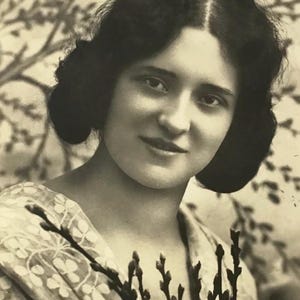 May include: A vintage black and white postcard showing a woman with dark hair in a classic updo, holding dark branches. The background features a blurred tree. The woman wears a light-coloured patterned shawl. The postcard has the number 1348.