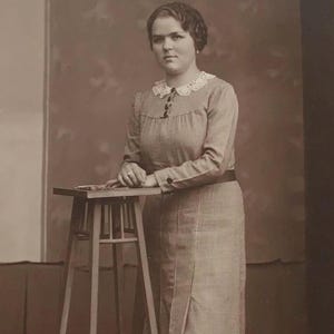 Puede incluir: Fotografía vintage en tonos sepia de una mujer con un vestido con cuello de encaje blanco, de pie junto a un pequeño taburete de madera. La mujer lleva un vestido con mangas largas y una falda con abertura. Lleva zapatos con correa.